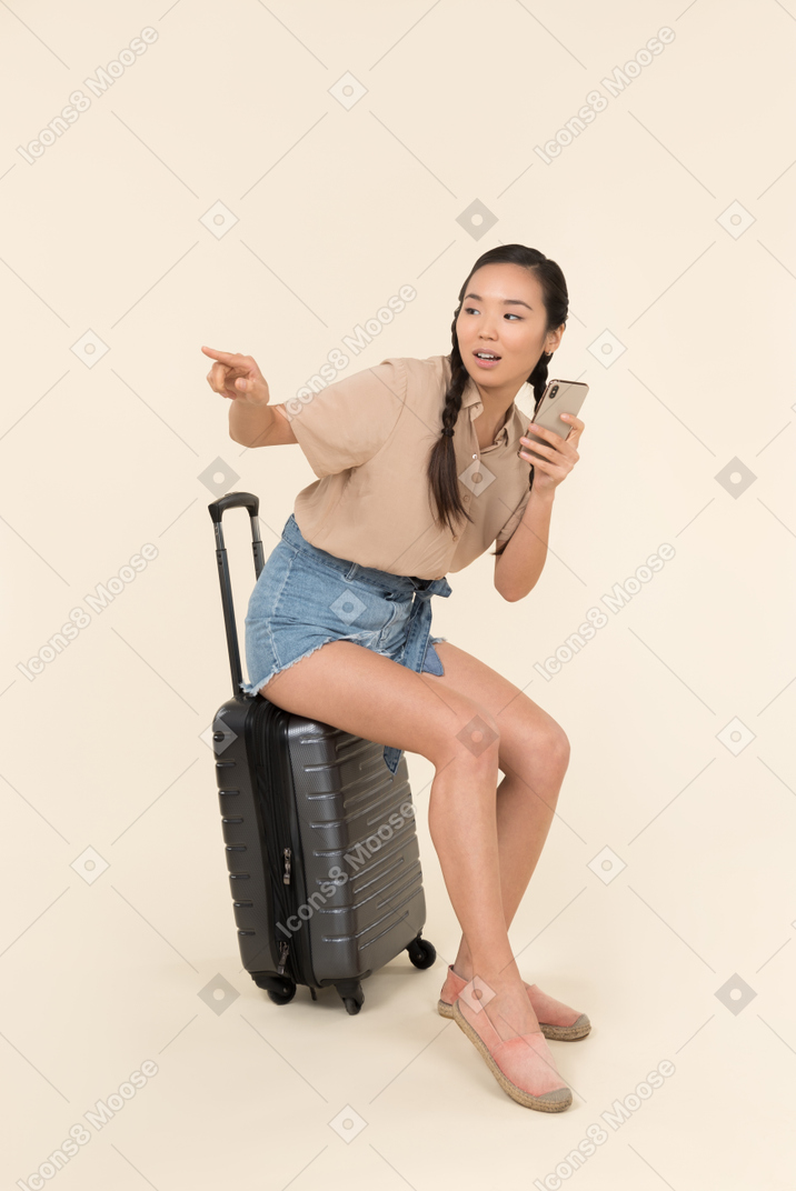 Young female traveller sitting on suitcase, holding phone and pointing aside