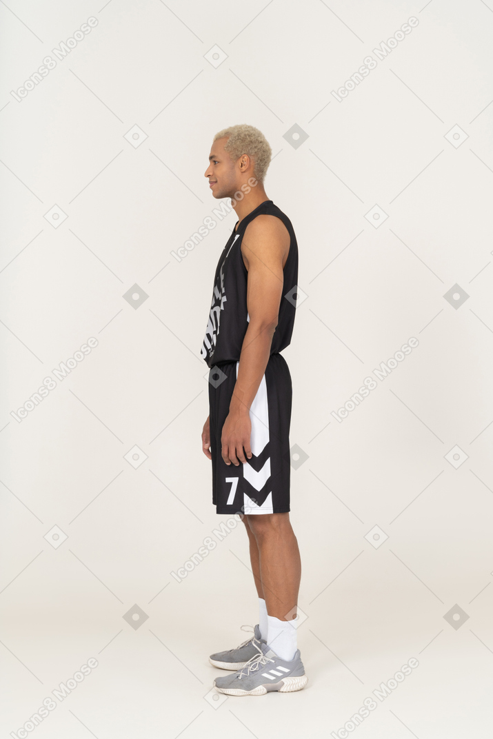 Vista lateral de un sonriente joven jugador de baloncesto de pie todavía