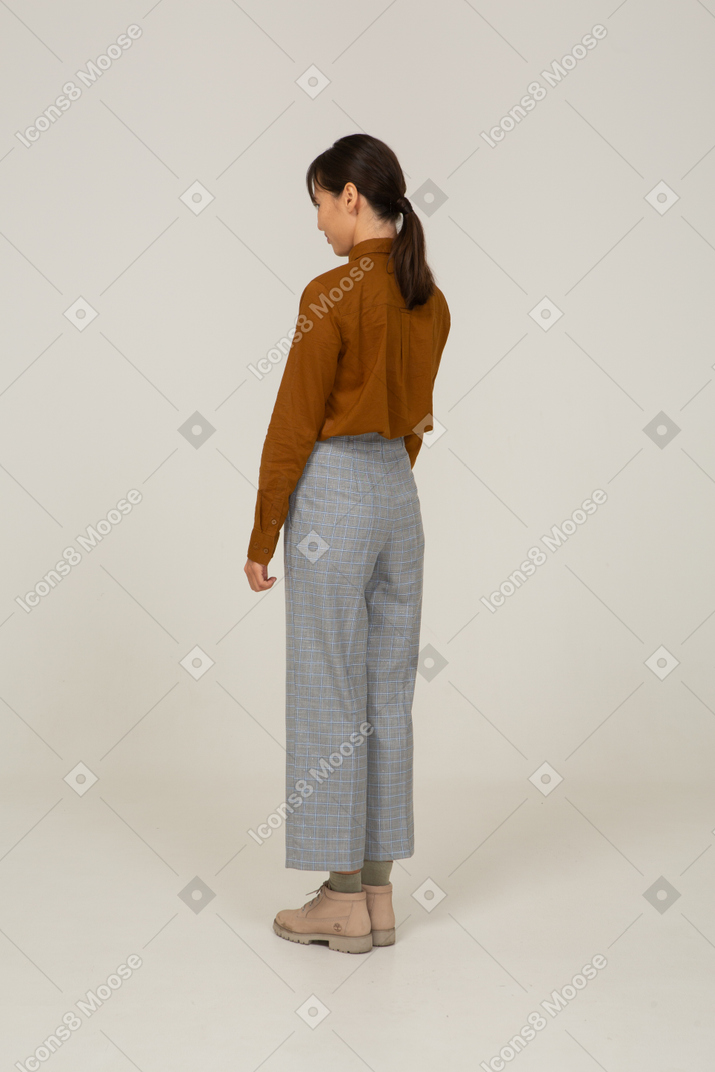 Vista traseira a três quartos de uma jovem mulher asiática de calça e blusa parada