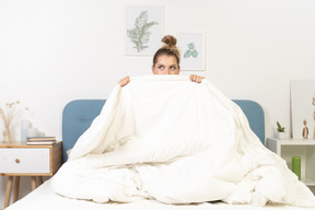 Vista frontal de una mujer joven en pijama escondido detrás de la manta en la cama