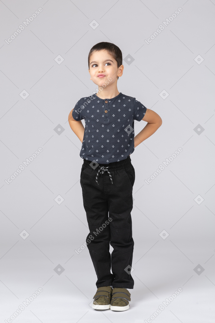 Вид спереди симпатичного мальчика в повседневной одежде, позирующего с руками на спине и корчащего рожи