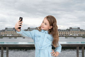 子供の女の子が川の背景の上に立って、selfieを作る