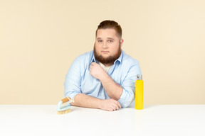 洗浄スプレーとブラシでテーブルに座っている深刻な探している若い太りすぎの人