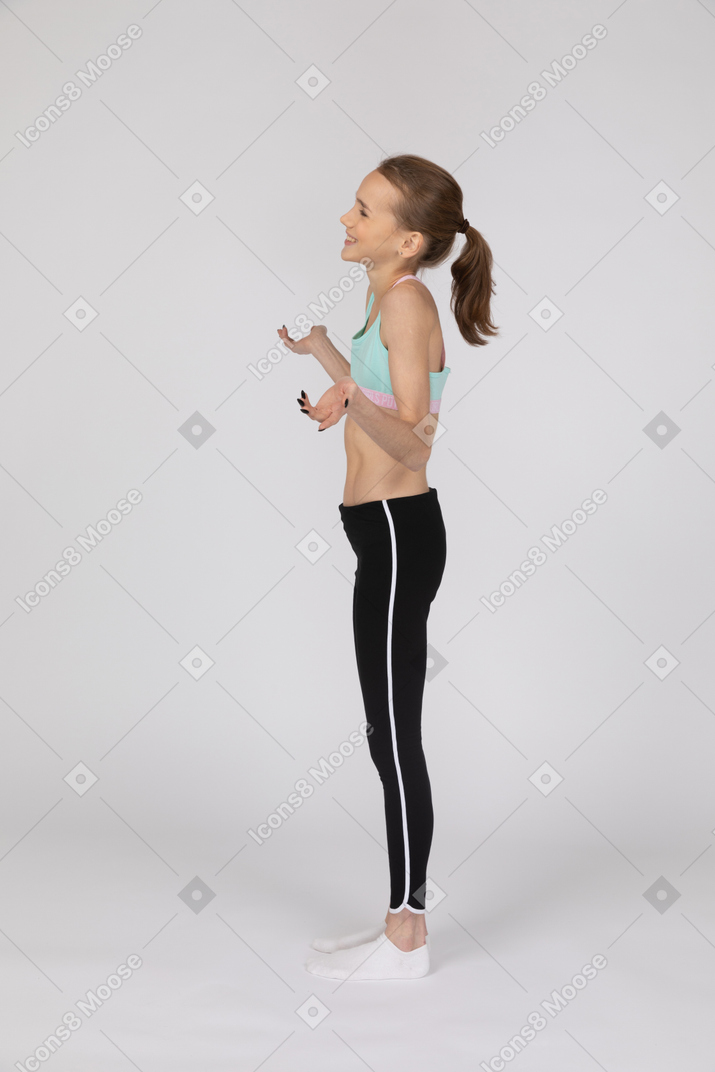 Vista lateral de una jovencita en ropa deportiva levantando las manos y sonriendo