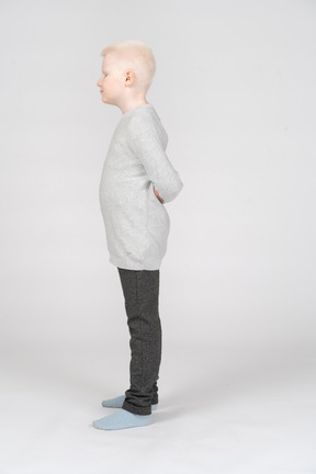 A blonde little boy in casual clothes standing and hiding hands behind