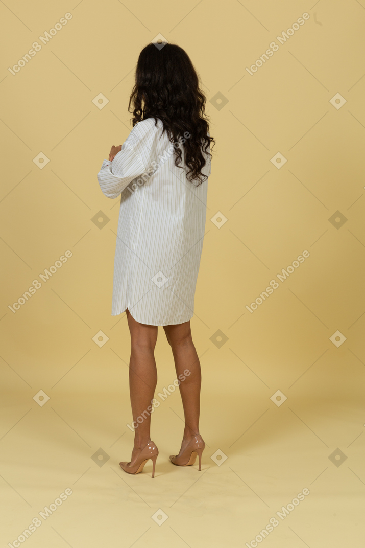 Three-quarter back view of a dark-skinned young female in white dress rolling up her sleeves