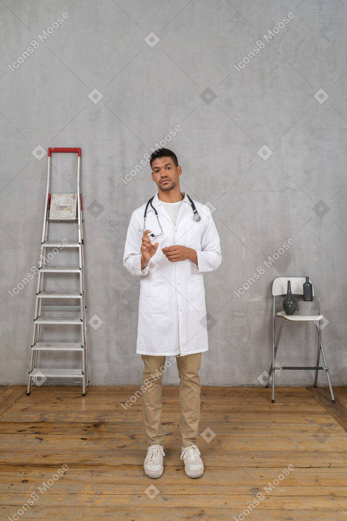 Vista frontale di un giovane dottore in piedi in una stanza con una scala e una sedia che mostra una dimensione di qualcosa