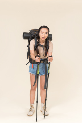 Tired young hiker woman standing with huge backpack and leaning on trekking poles