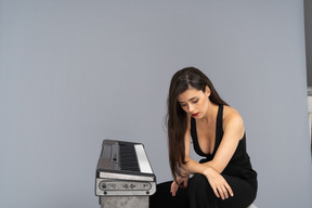 Full-length of a sad young lady in black sitting by the piano and holding hands on legs