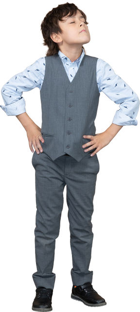 Вид спереди на симпатичного мальчика в сером костюме, позирующего с руками на бедрах и смотрящего вверх
