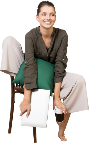 Front view of a smiling young woman sitting on a chair and holding her laptop & touching coffee cup
