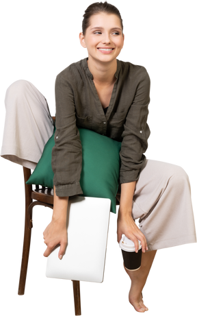 Vue de face d'une jeune femme souriante assise sur une chaise et tenant son ordinateur portable et touchant une tasse de café