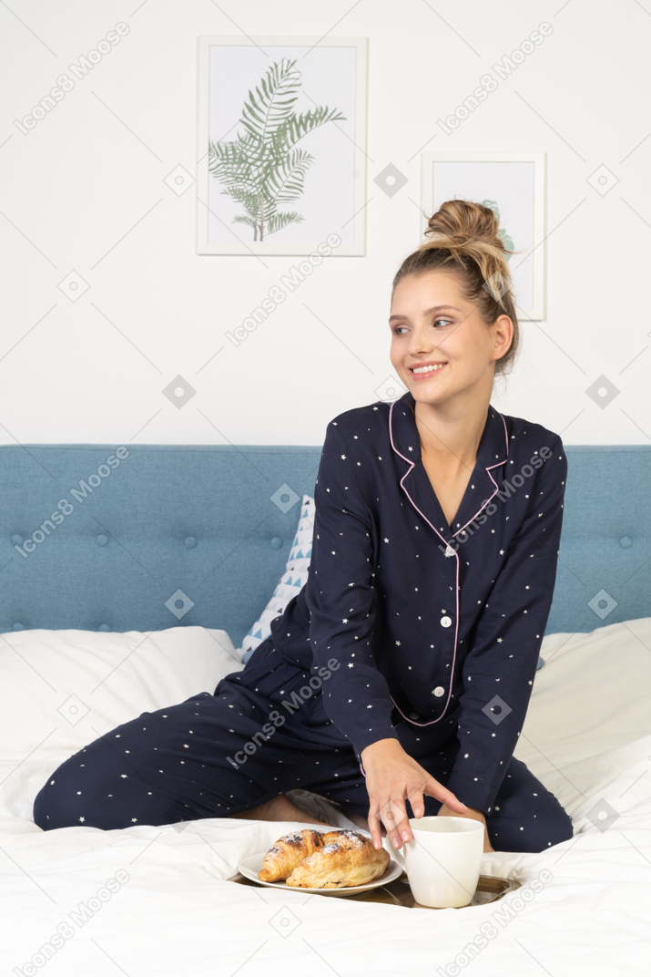 Vista frontal de uma jovem de pijama segurando uma xícara de café e alguns doces em uma bandeja