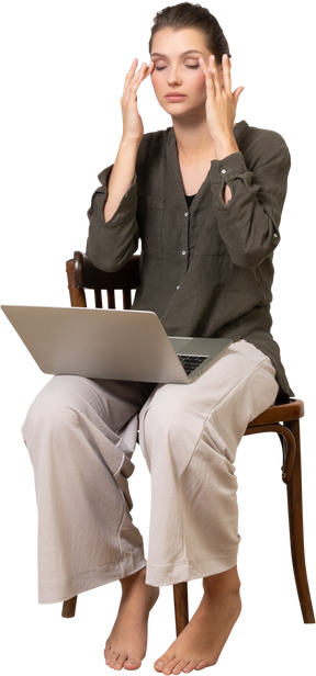 Vista de tres cuartos de una mujer joven ocupada con dolor de cabeza sentado en una silla con una computadora portátil