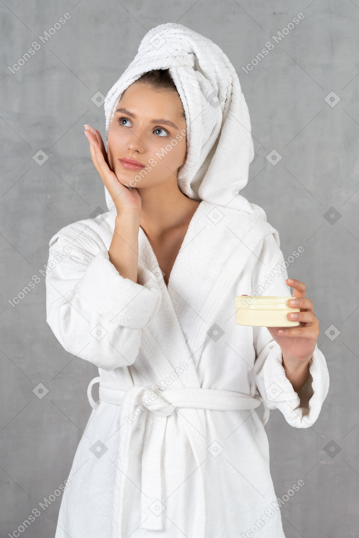 Woman in bathrobe touching her chin and holding tub of cream