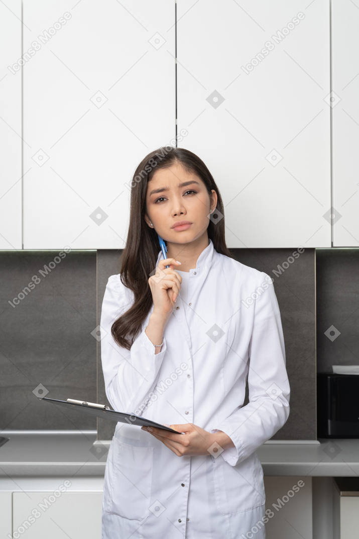 Vista frontal de una doctora perpleja sosteniendo un bolígrafo con una tableta y mirando a la cámara