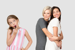 Two women hugging and a little girl putting lipstick on