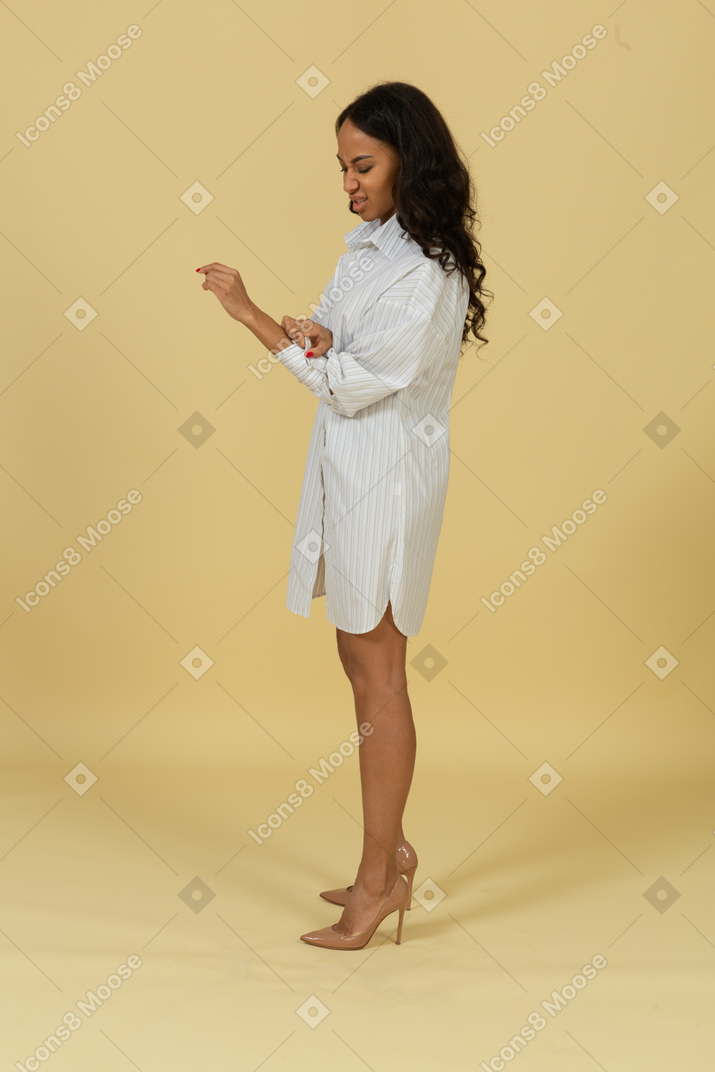 Вид сбоку на раздраженную смуглую девушку в белом платье, застегивающую рукава