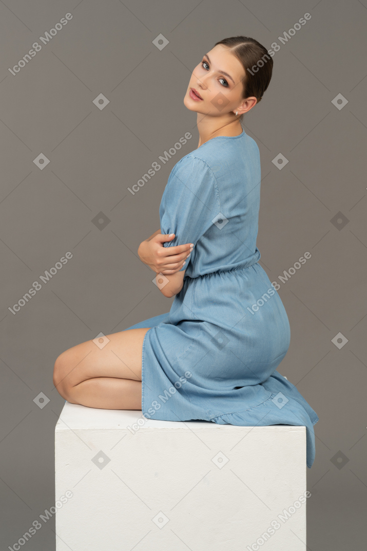 Vista posteriore della giovane donna in vestito blu che si siede sul cubo