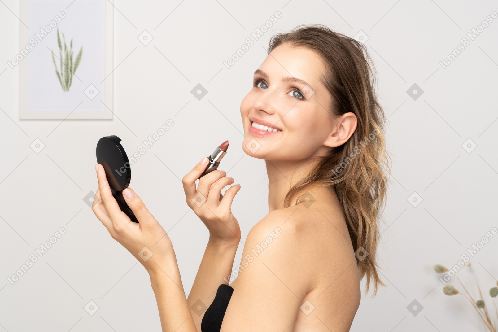 Side view of a smiling young woman applying lipstick while holding a mirror