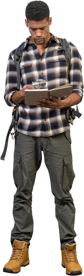 Vista frontal de un turista con una mochila examinando el bloc de notas a través de una lupa