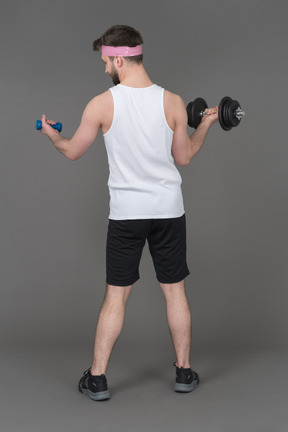 Slim young man holding heavy and light dumbbells back to camera