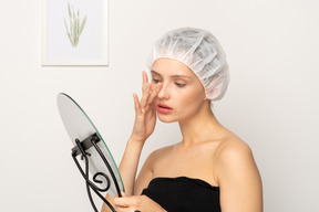 Young female patient in medical cap touching her nose while looking in the mirror
