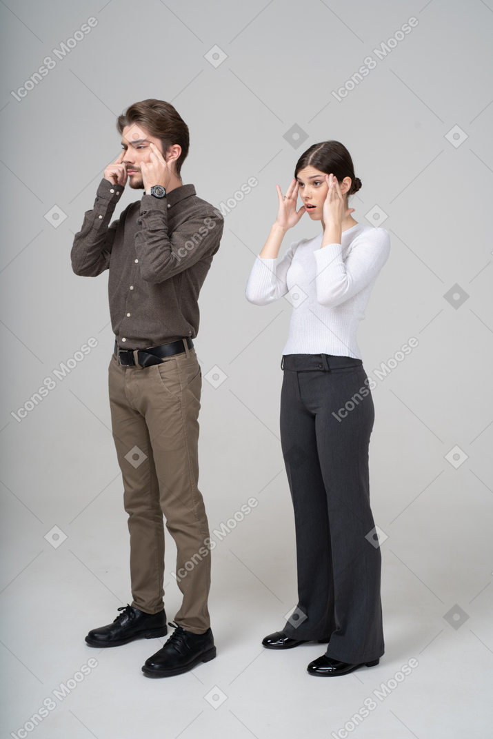 Three-quarter view of a young couple in office clothing touching face & narrowing eyes