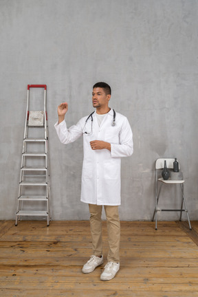 Three-quarter view of a young doctor standing in a room with ladder and chair showing a size of something