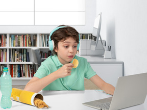 Boy in headphones sitting at the desk with laptop and eating chips