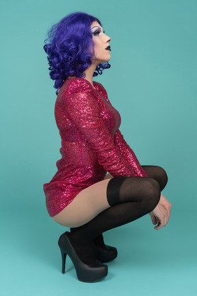 Side view of a drag queen in pink dress squatting with hands between knees