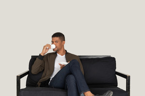 Vorderansicht eines jungen träumenden mannes, der auf einem sofa sitzt, während er kaffee trinkt