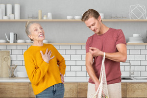 An older woman standing and laughing next to a man holding reusable bag in the kitchen