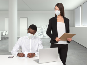 A woman wearing a face mask standing next to a man working on a laptop