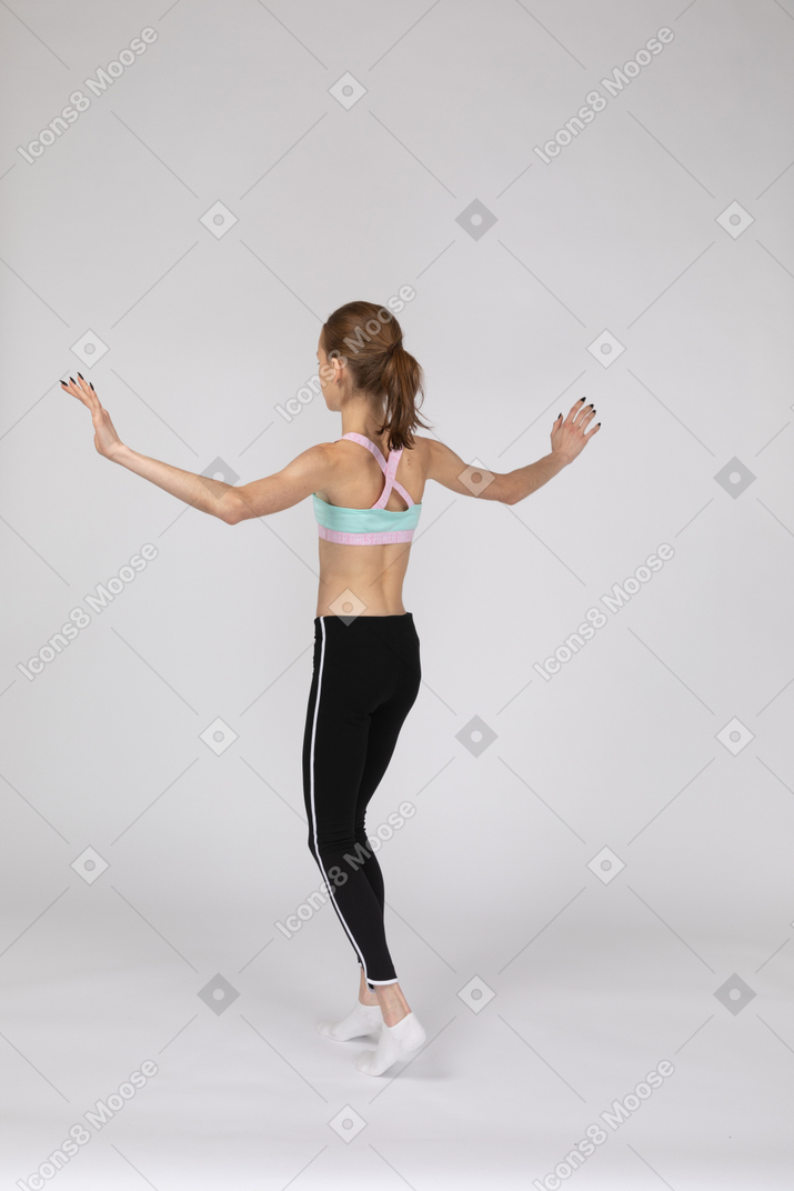 Три четверти сзади девушки-подростка в спортивной одежде, балансирующей на цыпочках и поднимающей руки