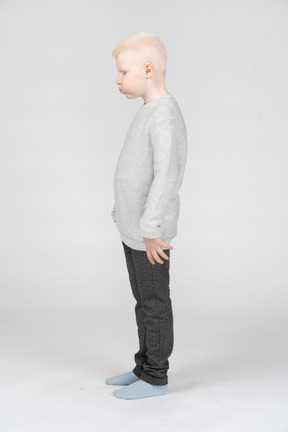 Vista lateral de un niño chico rubio en ropa casual soplando las mejillas mientras mira hacia abajo