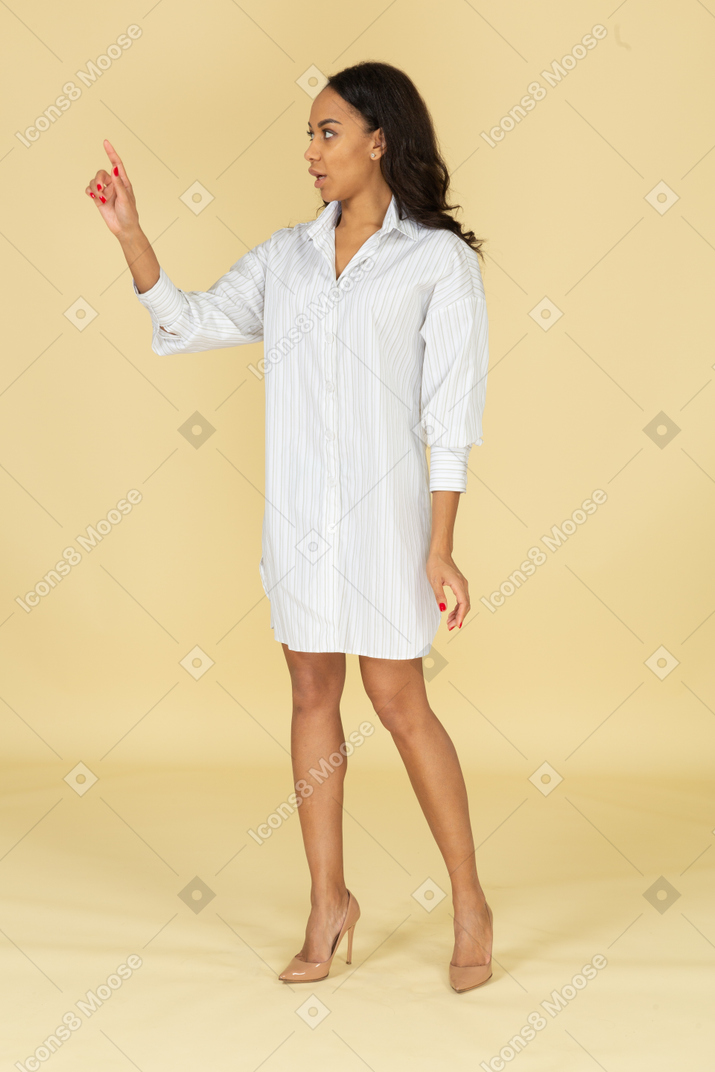 Three-quarter view of a dark-skinned young female in white dress raising hand while explaining something