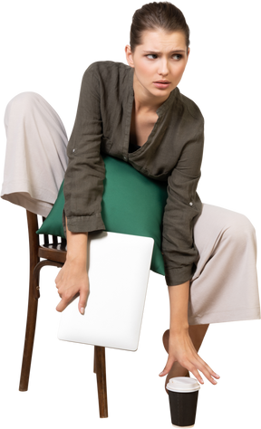 Vista frontal de una mujer joven confundida sentada en una silla y sosteniendo su computadora portátil y tocando la taza de café