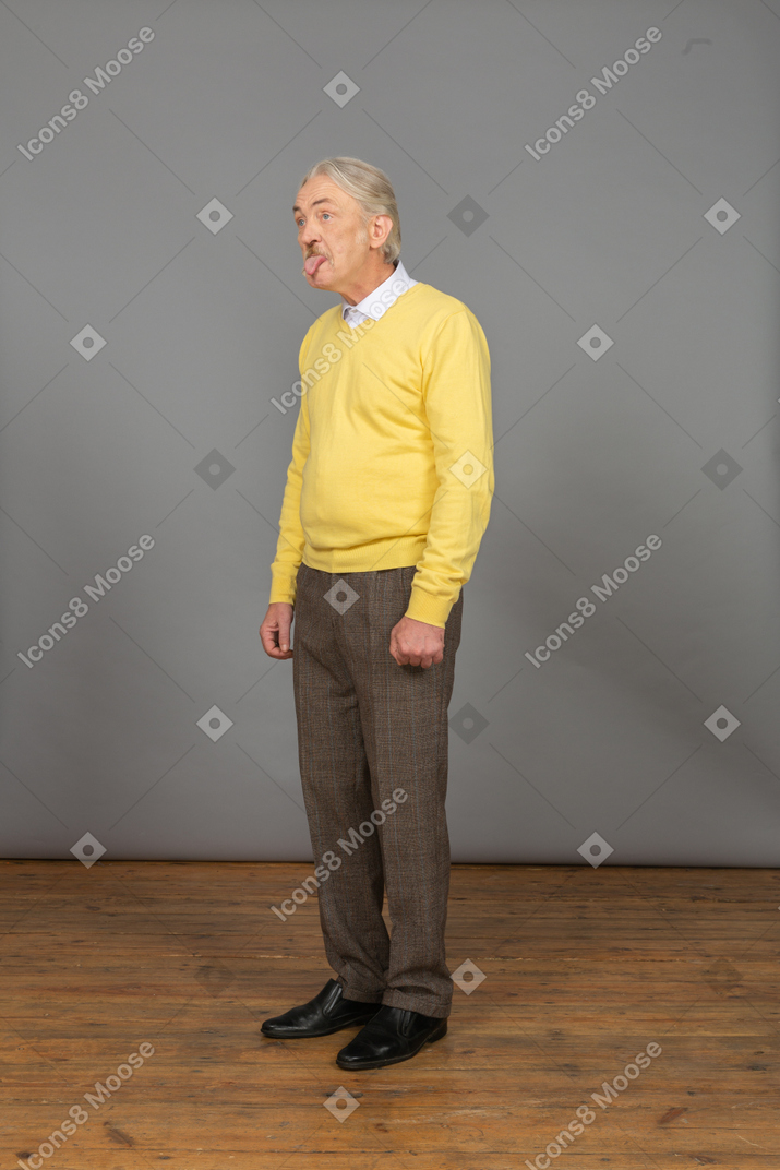 Vista de tres cuartos de un anciano curioso en jersey amarillo mostrando la lengua y mirando a un lado