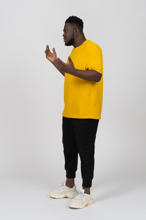 노란색 티셔츠를 입고 몸짓으로 말다툼을 하는 검은 피부의 젊은 남자의 3/4 보기