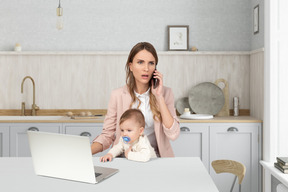 A woman sitting at a kitchen table talking on a cell phone while holding a baby