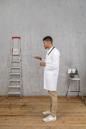 Vista lateral de un joven médico de pie en una habitación con escalera y silla mostrando el tamaño de algo