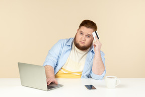 Pensieroso giovane uomo in sovrappeso seduto davanti al computer portatile e fare shopping online