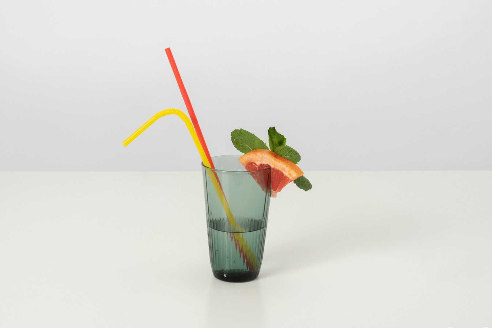 A glass of cold water with a slice of grapefruit and some mint leaves on it, with two colorful plastic straws because drinking from both at the same time is double fun