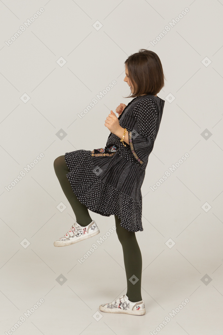 Vista laterale di una bambina felice in abito stringendo i pugni e alzando la gamba