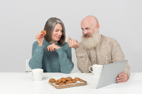 Couple âgé assis à la table, ayant des biscuits et du café et regardant un film sur une tablette