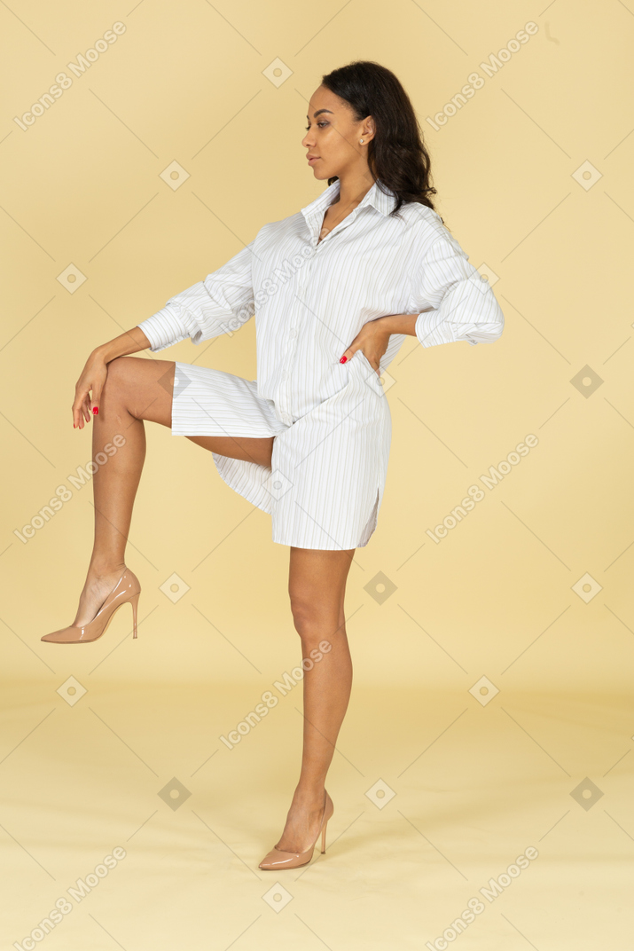 Vista di tre quarti di una giovane donna dalla carnagione scura in abito bianco alzando la gamba