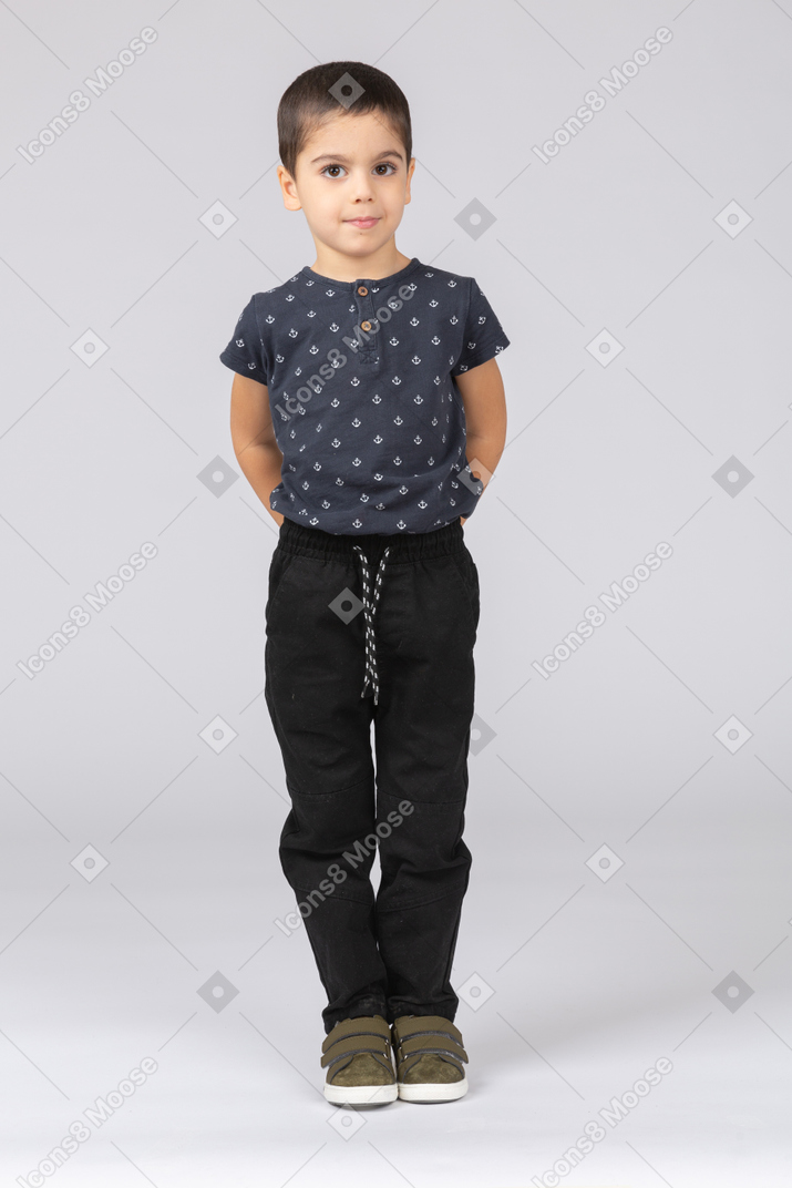 Front view of a cute boy standing with hands behind back and looking at camera
