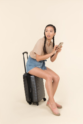 Young female traveller sitting on suitcase, holding phone and pointing aside