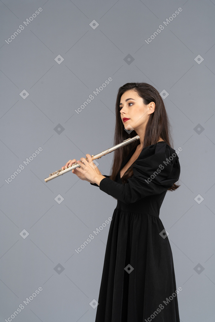 Вид сбоку серьезной молодой леди в черном платье с флейтой